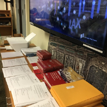 Photo of desk below mounted flat screen featuring an LAX desktop wallpaper, the desk contains a stack of manila folders as well as the varies needed applications and pamphlets .